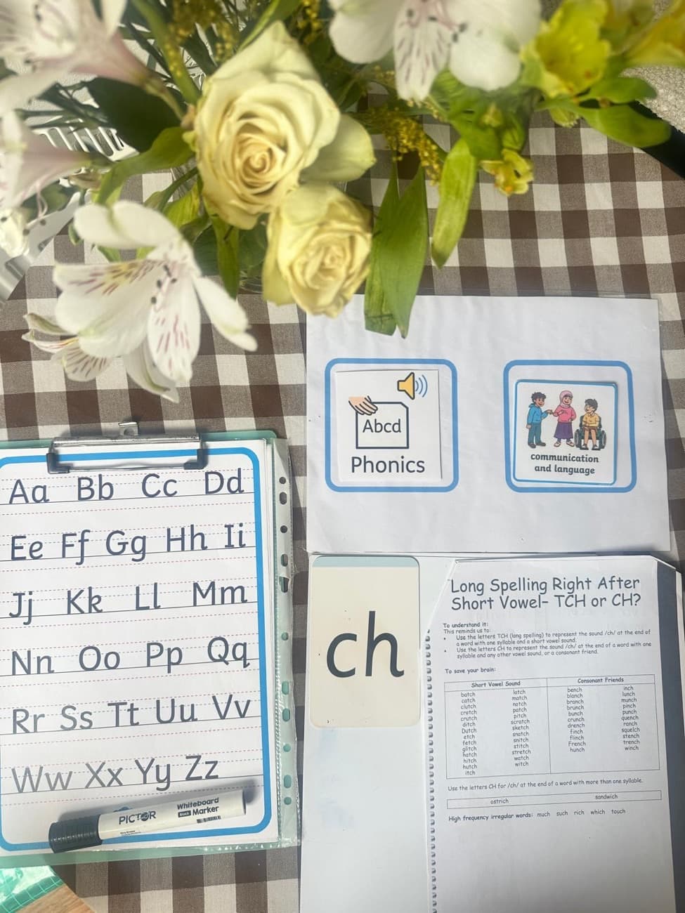 Phonics teaching resources on a kitchen table: an alphabet board, picture cards for phonics and communication, a 'ch' sound card and a spelling reference sheet.